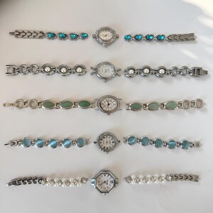 May include: Five vintage women's watches with silver-toned metal straps. Each watch has a unique design, with turquoise, pearl, and jade-coloured embellishments. The watch faces display Roman numerals and decorative elements.