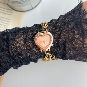 Puede incluir: Un reloj dorado en forma de coraz&oacute;n con una esfera color melocot&oacute;n y peque&ntilde;os detalles de cristal. El reloj est&aacute; sujeto a una delicada cadena dorada. El reloj est&aacute; sobre una manga de encaje negro.