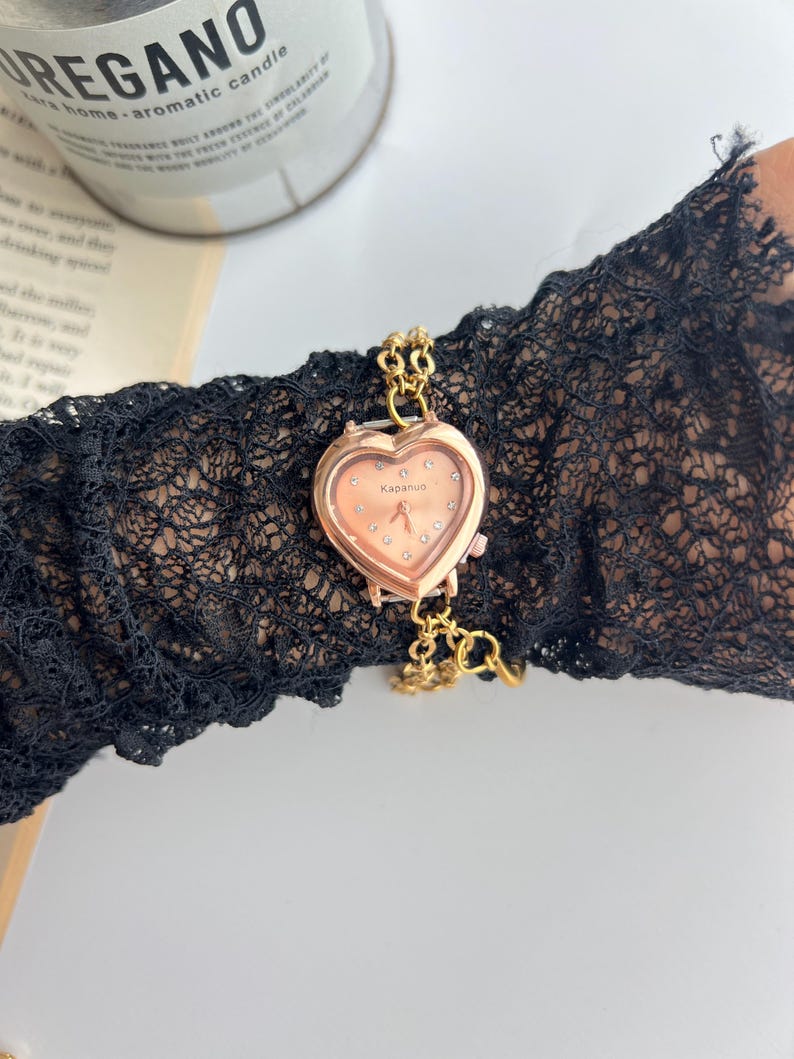 Puede incluir: Un reloj dorado en forma de coraz&oacute;n con una esfera color melocot&oacute;n y peque&ntilde;os detalles de cristal. El reloj est&aacute; sujeto a una delicada cadena dorada. El reloj est&aacute; sobre una manga de encaje negro.