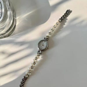 May include: Silver wristwatch with a white face and a bracelet of pearls and rhinestones. The watch has an oval face and a delicate design, suitable for everyday wear or special occasions.