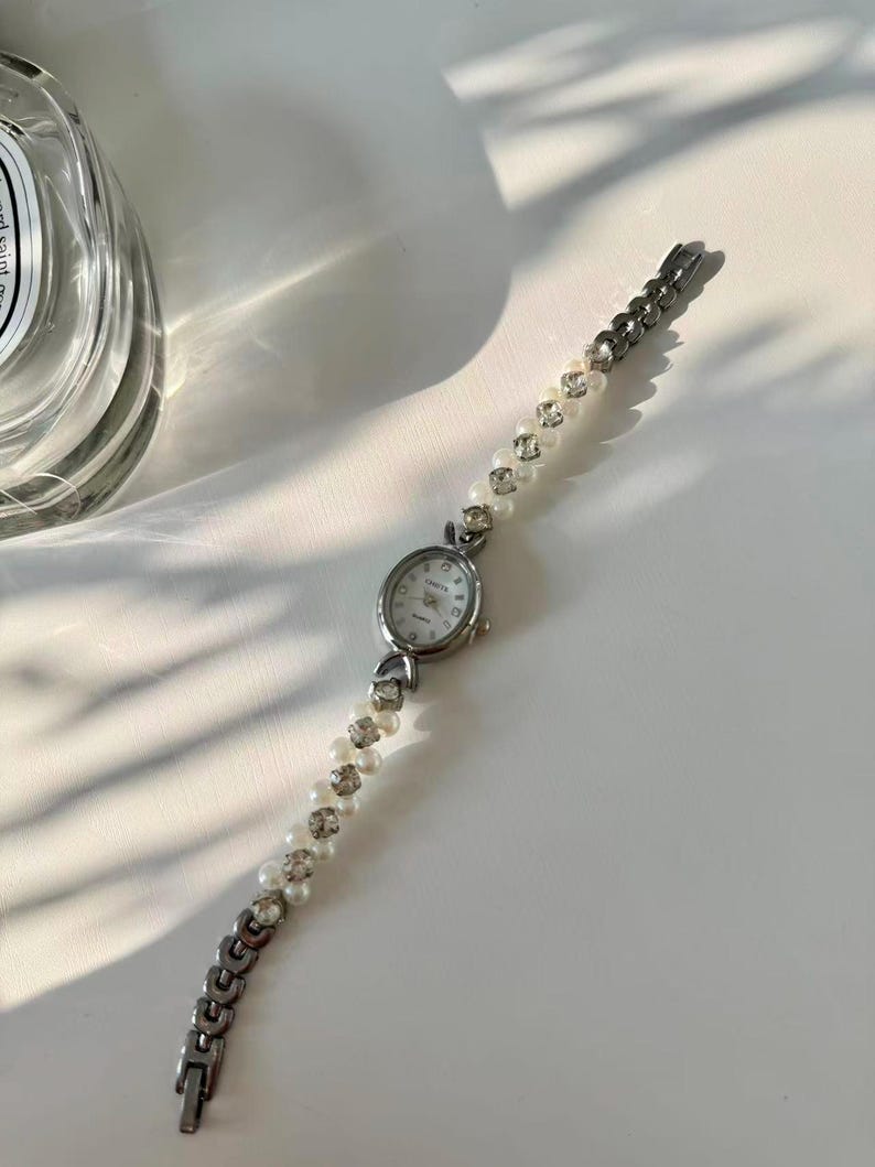 May include: Silver wristwatch with a white face and a bracelet of pearls and rhinestones. The watch has an oval face and a delicate design, suitable for everyday wear or special occasions.