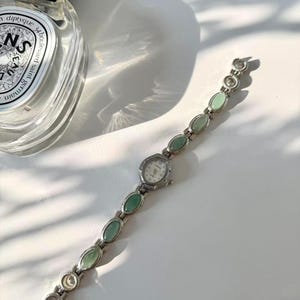 May include: A silver wristwatch with a white face and a band adorned with oval, pale green stones. The watch is displayed on a white surface, with a perfume bottle partially visible in the top left corner.
