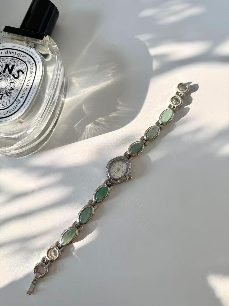 May include: A silver wristwatch with a white face and a band adorned with oval, pale green stones. The watch is displayed on a white surface, with a perfume bottle partially visible in the top left corner.