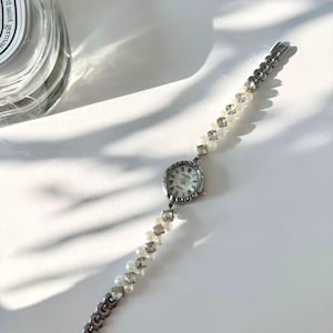 May include: A silver wristwatch with a round face and a white dial. The watch strap combines pearls, crystals, and silver links. The watch has a delicate, feminine design. The watch is suitable for a small wrist.