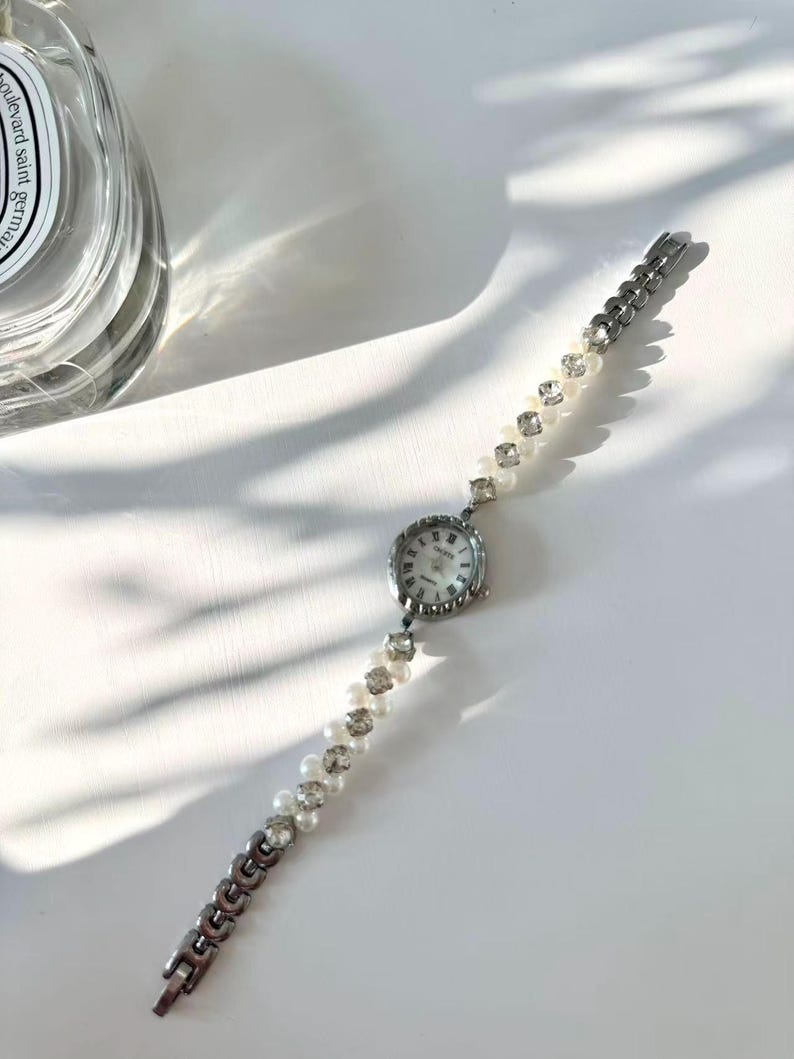 May include: A silver wristwatch with a round face and a white dial. The watch strap combines pearls, crystals, and silver links. The watch has a delicate, feminine design. The watch is suitable for a small wrist.