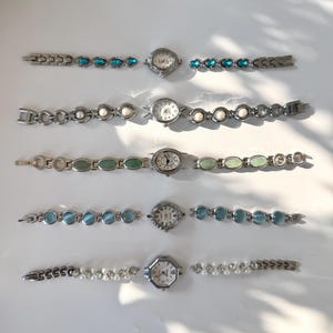 May include: Five silver-coloured wristwatches with decorative straps. The straps feature turquoise, white pearl, green, blue, and pearl embellishments. The watch faces are round or octagonal, with visible hands and numbers.