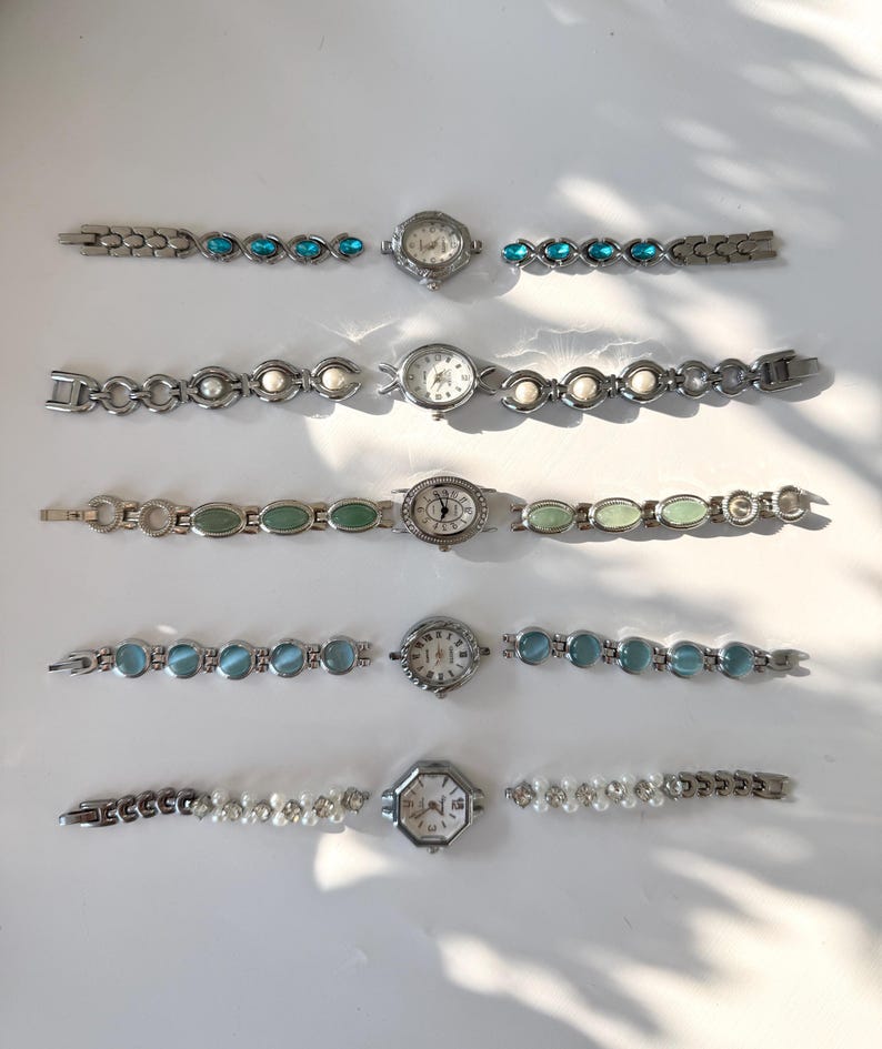 May include: Five silver-coloured wristwatches with decorative straps. The straps feature turquoise, white pearl, green, blue, and pearl embellishments. The watch faces are round or octagonal, with visible hands and numbers.