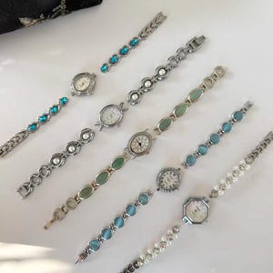 May include: A selection of silver-coloured wristwatches with decorative straps. The straps are adorned with blue, green, and white gemstones, and pearls. The watch faces are round and octagonal with black numerals and hands. A black floral pouch is in the background.