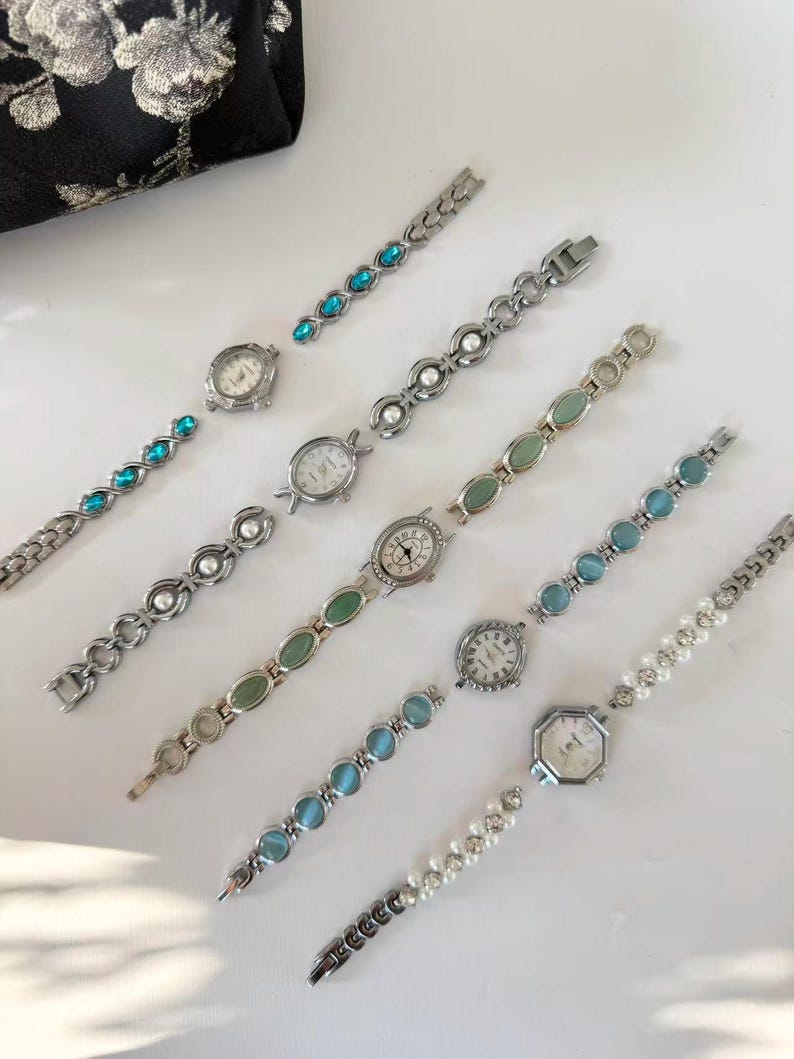 May include: A selection of silver-coloured wristwatches with decorative straps. The straps are adorned with blue, green, and white gemstones, and pearls. The watch faces are round and octagonal with black numerals and hands. A black floral pouch is in the background.