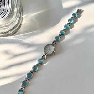 May include: A silver wristwatch featuring a white watch face and a strap embellished with light blue, circular stones. The watch is presented on a white surface, with a glass bottle visible in the background.