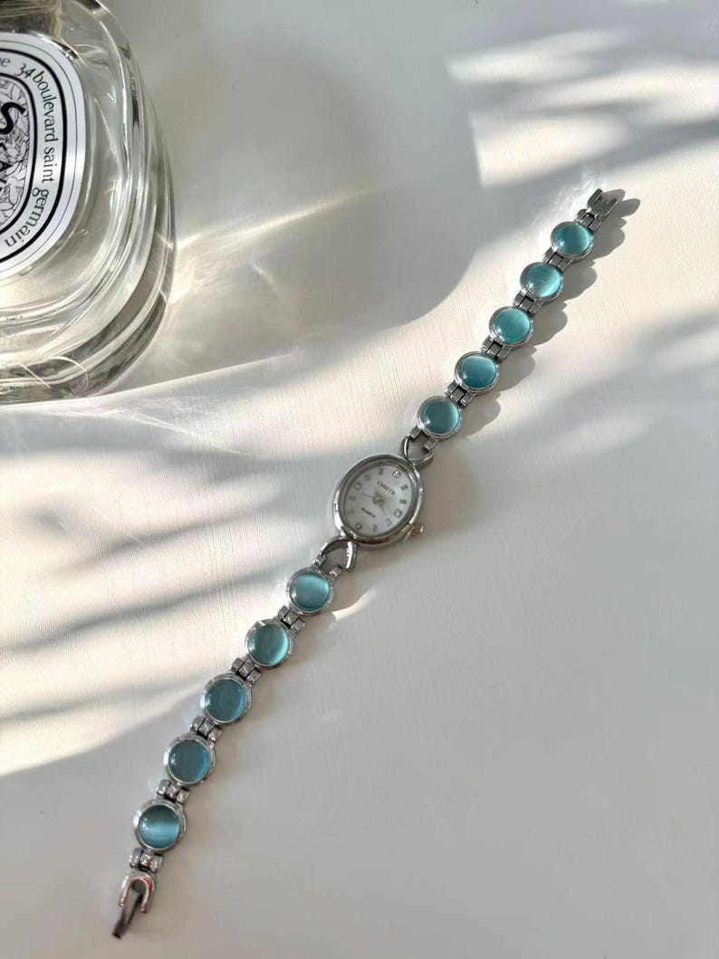 May include: A silver wristwatch featuring a white watch face and a strap embellished with light blue, circular stones. The watch is presented on a white surface, with a glass bottle visible in the background.