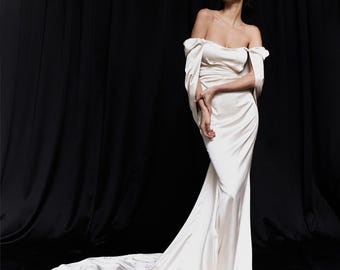 May include: An off-white, off-the-shoulder gown with a flowing train. The dress features draped fabric and sleeves that extend from the shoulders. The model is standing in front of a black backdrop.