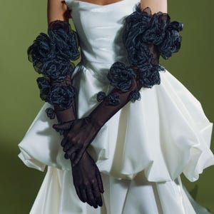 May include: A white strapless dress with a bubble skirt, paired with sheer black gloves adorned with large, dark floral accents. The gloves extend to the elbows, and the flowers are clustered on the upper arms and hands.