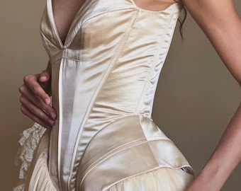 May include: A strapless champagne-coloured satin corset bodice featuring a deep V-neckline and structured paneling, paired with a layered tulle skirt that includes delicate white lace trim detailing along the side hip area.
