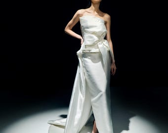 Puede incluir: Un vestido de satén blanco sin tirantes con un lazo en la cintura y una cola fluida. El vestido es largo hasta el suelo y tiene un diseño elegante. La modelo lleva tacones negros.
