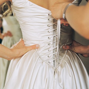Puede incluir: Un vestido de novia marfil sin tirantes con espalda de cors&eacute; que se est&aacute; atando. La tela de sat&eacute;n es lisa y el corpi&ntilde;o est&aacute; intrincadamente detallado. El vestido est&aacute; siendo ajustado por varias manos.