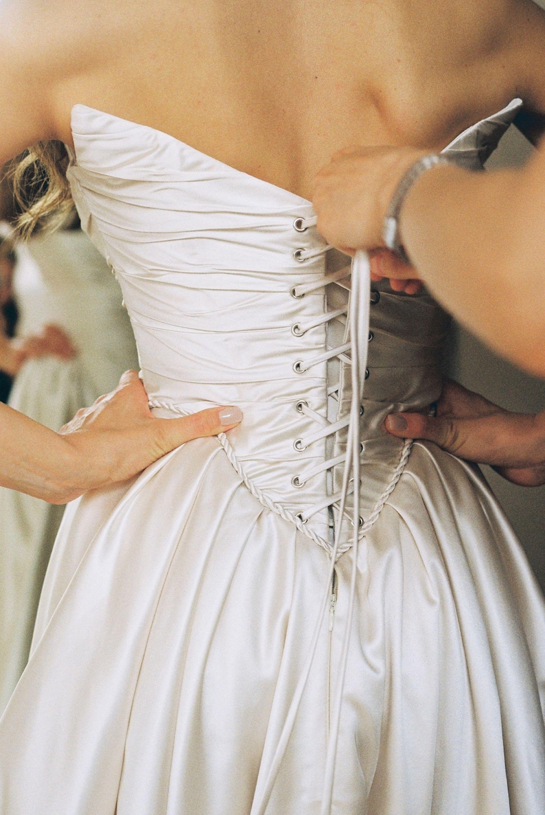 Puede incluir: Un vestido de novia marfil sin tirantes con espalda de cors&eacute; que se est&aacute; atando. La tela de sat&eacute;n es lisa y el corpi&ntilde;o est&aacute; intrincadamente detallado. El vestido est&aacute; siendo ajustado por varias manos.