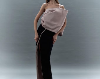 May include: A formal outfit featuring a blush-colored, one-shoulder top with a draped design. The top is paired with a black, floor-length skirt with a high slit revealing a contrasting taupe panel. The outfit is elegant and modern.