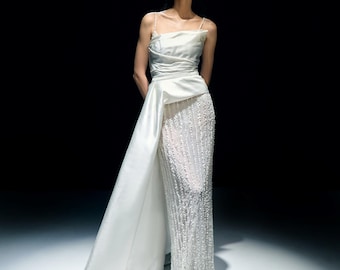 Puede incluir: Un vestido de noche blanco con un corpiño drapeado y una falda fluida. La falda está adornada con hileras verticales de pequeñas cuentas brillantes. Una larga y elegante cola se extiende desde la cintura, añadiendo un toque dramático al diseño.