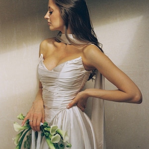 Puede incluir: Vestido de novia de sat&eacute;n color crema con corpi&ntilde;o drapeado y falda amplia. El vestido presenta un escote coraz&oacute;n y detalles de cuerda. La novia sostiene un ramo de calas blancas. Un elegante vestido de novia.