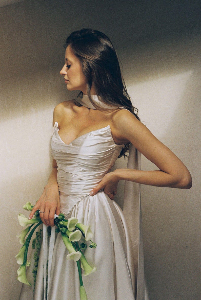 Puede incluir: Vestido de novia de sat&eacute;n color crema con corpi&ntilde;o drapeado y falda amplia. El vestido presenta un escote coraz&oacute;n y detalles de cuerda. La novia sostiene un ramo de calas blancas. Un elegante vestido de novia.