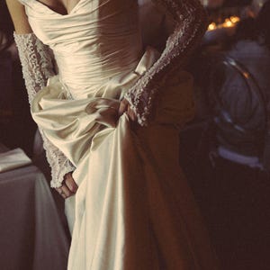 May include: A cream-colored satin wedding gown with a sweetheart neckline and a draped bodice. The gown features a full skirt and is paired with lace gloves. The overall style is elegant and vintage-inspired, suitable for a formal event.