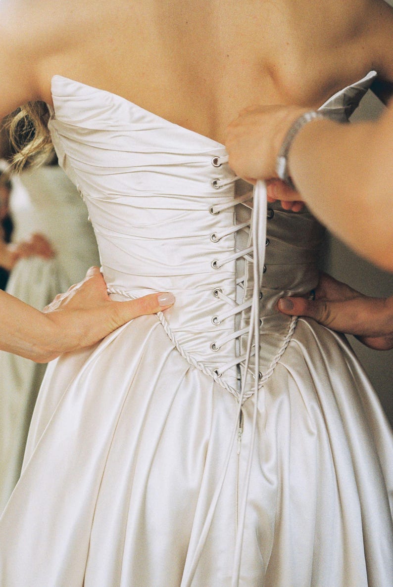 May include: A strapless ivory wedding dress with a corset back. The dress is being laced up by two people. The satin fabric has a subtle sheen, and the bodice features delicate pleating.