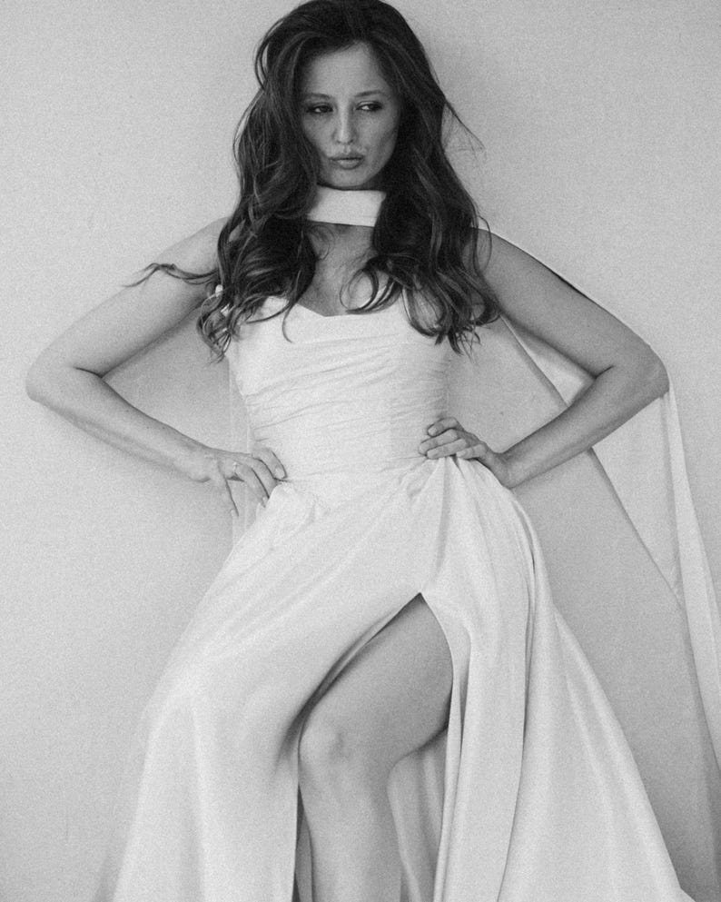 Puede incluir: Fotograf&iacute;a en blanco y negro de una mujer con un vestido blanco con una abertura alta y una capa. El vestido tiene un escote coraz&oacute;n y est&aacute; ajustado a la cintura. La mujer tiene el pelo largo y ondulado y posa con las manos en las caderas.