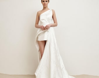 May include: A white, one-shoulder wedding dress with a short skirt and a long train. The dress has a textured fabric and a fitted bodice. The model is standing against a neutral background.