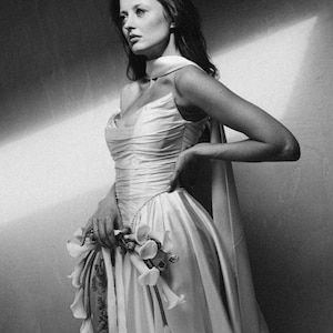 Puede incluir: Fotograf&iacute;a en blanco y negro de una mujer con un vestido formal. El vestido es de color claro, con un corpi&ntilde;o drapeado y una falda amplia. Sostiene un ramo de flores. La imagen es un retrato, con la mujer mirando hacia un lado.
