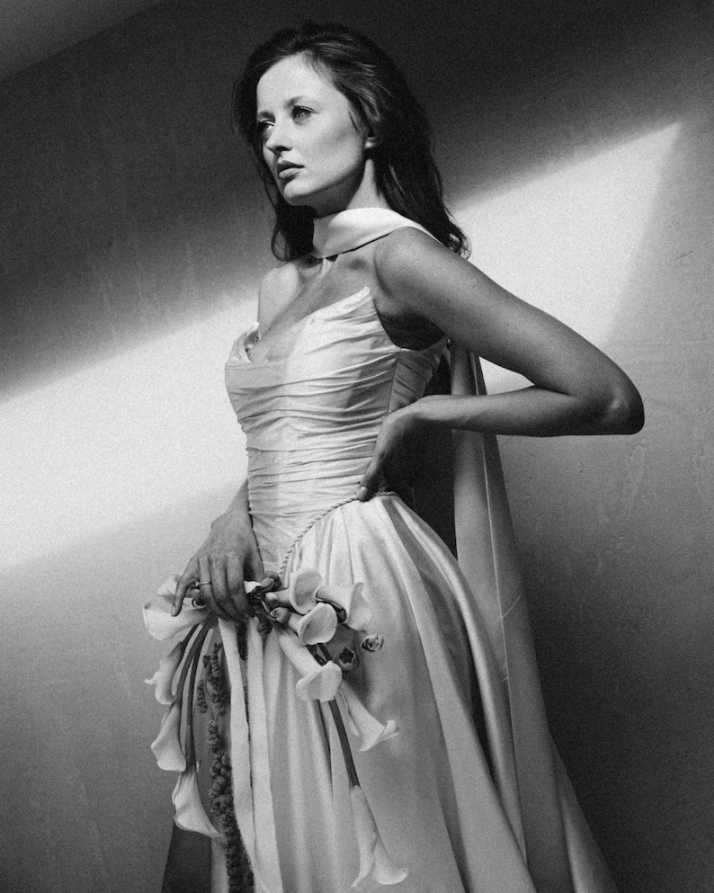 Puede incluir: Fotograf&iacute;a en blanco y negro de una mujer con un vestido formal. El vestido es de color claro, con un corpi&ntilde;o drapeado y una falda amplia. Sostiene un ramo de flores. La imagen es un retrato, con la mujer mirando hacia un lado.