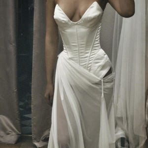 May include: A white corset-style bodice featuring a structured, paneled design with a deep V-neckline. The garment is paired with a sheer, flowing skirt overlay that drapes from the waist, creating a layered effect with visible vertical seams.