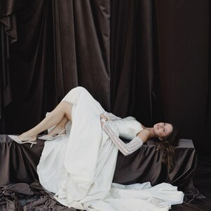 May include: A woman wearing a white wedding dress with long sleeves and a flowing skirt. She is lying on a brown surface with a dark brown backdrop. The dress has a fitted bodice and a long train. She is wearing white heels.
