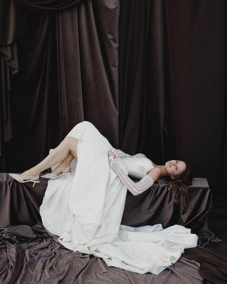 May include: A woman wearing a white wedding dress with long sleeves and a flowing skirt. She is lying on a brown surface with a dark brown backdrop. The dress has a fitted bodice and a long train. She is wearing white heels.