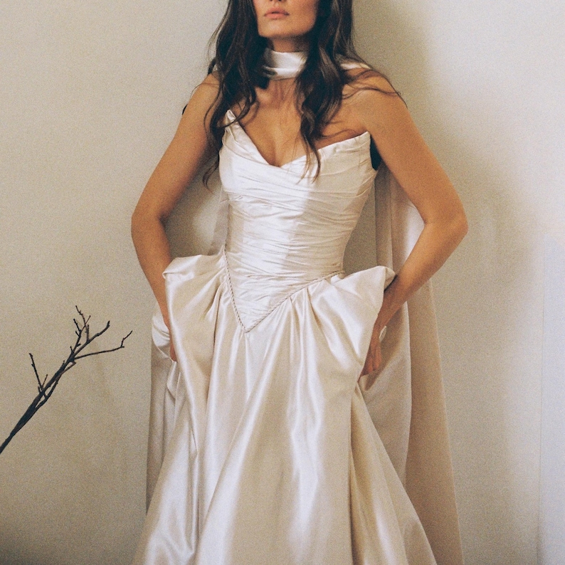 Puede incluir: Un vestido de sat&eacute;n color crema con escote coraz&oacute;n y falda amplia. El vestido presenta un corpi&ntilde;o ajustado con fruncidos y una capa que cae sobre los hombros. Una delicada cadena adorna el corpi&ntilde;o.