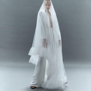 May include: A white bridal ensemble featuring a blazer, wide-leg pants, and a sheer veil. The veil cascades down the front and back, creating a flowing silhouette. The outfit is simple yet elegant, perfect for a wedding.