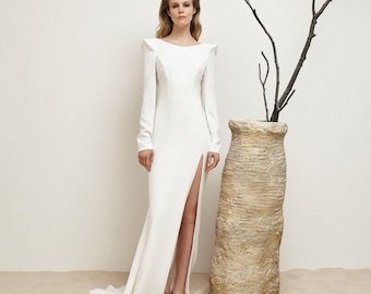 May include: A white, long-sleeved gown with a high slit and a train. The dress features structured shoulders and a fitted bodice. A tall, textured vase with a branch is next to the dress.