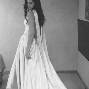 Puede incluir: Imagen en escala de grises de un vestido formal. El vestido es de color claro, posiblemente marfil o blanco, con un corpi&ntilde;o ajustado y una falda fluida que se arrastra por detr&aacute;s. El vestido tiene un detalle de faja larga y drapeada. La persona est&aacute; mirando por encima del hombro.