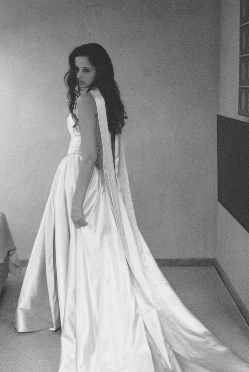 Puede incluir: Imagen en escala de grises de un vestido formal. El vestido es de color claro, posiblemente marfil o blanco, con un corpi&ntilde;o ajustado y una falda fluida que se arrastra por detr&aacute;s. El vestido tiene un detalle de faja larga y drapeada. La persona est&aacute; mirando por encima del hombro.