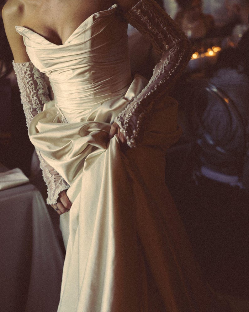 Puede incluir: Un vestido de novia color crema con un corpi&ntilde;o drapeado y una falda amplia. El vestido presenta mangas largas de encaje. La novia sostiene la falda del vestido. El tema general es la moda nupcial.