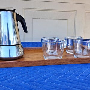 May include: A stainless steel stovetop espresso maker with a black handle sits on a rectangular wooden serving tray alongside three clear glass cups. The tray has angled corners and a smooth finish, providing a base for the coffee set.