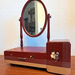 May include: A dark red wooden vanity mirror with an oval glass pane supported by two vertical posts. The base features a horizontal drawer with a metal handle and a side compartment decorated with a small illustration of a figure and white lines.