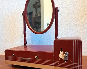 May include: A dark red wooden vanity mirror with an oval glass pane supported by two vertical posts. The base features a horizontal drawer with a metal handle and a side compartment decorated with a small illustration of a figure and white lines.