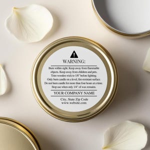 May include: A gold-colored candle tin with a white label displaying a warning and company information. The label includes safety instructions for use. White flower petals are scattered around the tin.