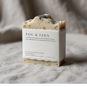 May include: A handcrafted soap bar with a white label reading "FOG & FERN". The soap is a light beige colour, speckled with visible flecks and topped with dried herbs. The label also reads "HANDCRAFTED SOAP" and "4.5 OZ (127 g)".