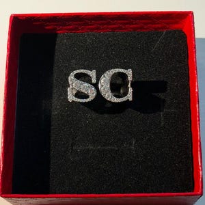 Puede incluir: Broche plateado con las letras "SC" incrustadas con pequeñas piedras preciosas transparentes, presentado en una caja roja con interior negro. El broche es el punto focal, con la caja abierta para mostrarlo.