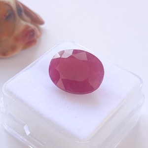 Puede incluir: Una gema de rubí facetada, de forma ovalada, exhibida sobre una superficie blanca dentro de una caja de plástico transparente. El rubí exhibe un tono rojo intenso, con múltiples facetas que capturan la luz.