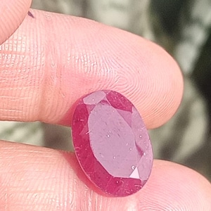 May include: A faceted, oval-shaped ruby gemstone. The ruby is a deep, rich red color, with multiple facets that catch the light. The gemstone is held between two fingers, with a blurred background.