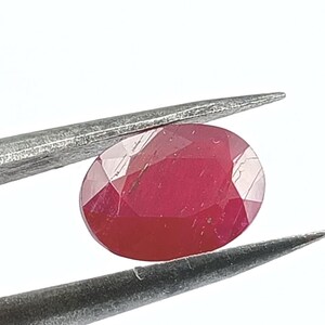 May include: A faceted, oval-shaped ruby gemstone, held by silver tweezers. The ruby displays a deep red color with visible internal facets, reflecting light. The background is plain white, emphasizing the gem's clarity and color.