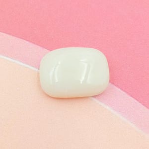 Puede incluir: Una piedra lisa, de color blanquecino, de forma ovalada, posiblemente una gema pulida o un elemento decorativo. La piedra tiene una superficie brillante y está colocada sobre un fondo de tela rosa y melocotón.